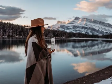 Woman in a wide-brimmed hat standing at the edge of a still mountain lake with snowy peaks reflected