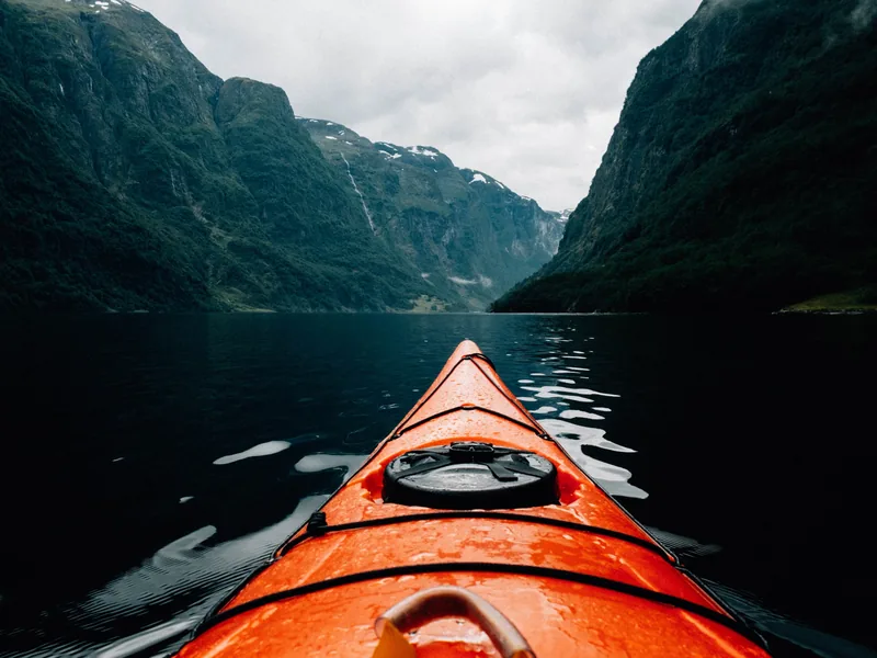 Bow of an orange kayak on dark fjord water between steep green mountains