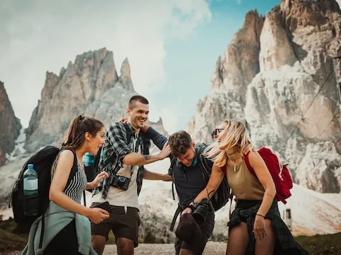 Four hikers with backpacks laughing together on a rocky trail below jagged Dolomite peaks