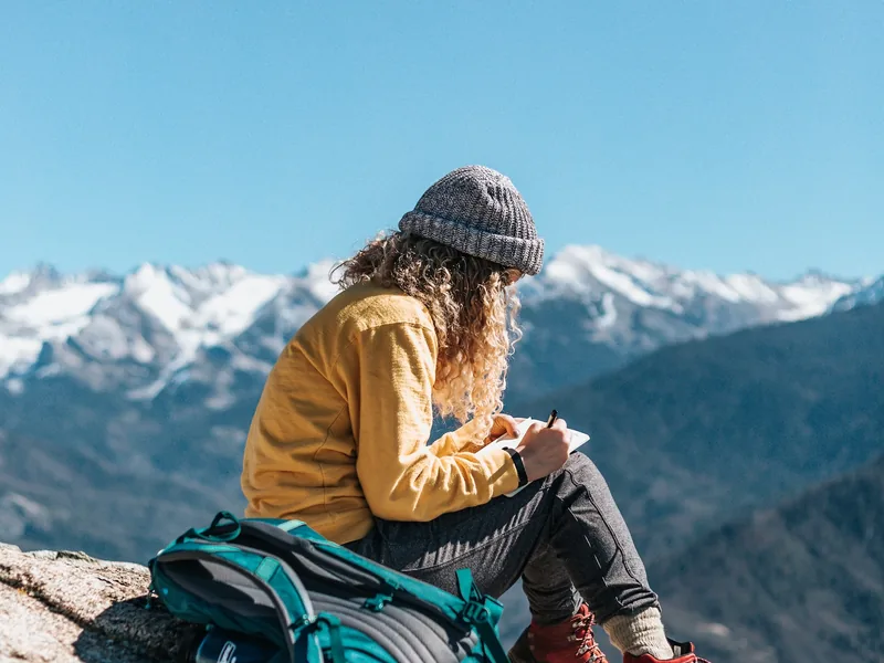 Hiker sitting on a rocky ridge with a teal backpack, snow-capped mountains behind
