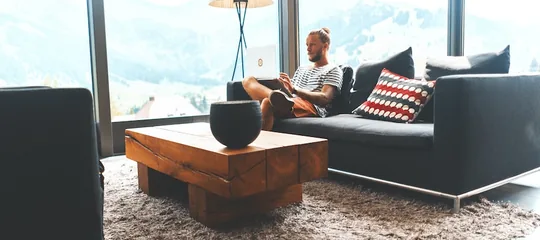 Man reading on a sofa in a living room with floor-to-ceiling windows